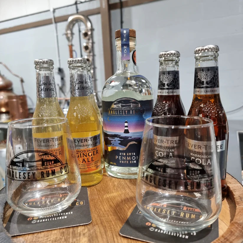 Collection of bottles and glasses with Anglesey Rum Co branding on a wooden surface.