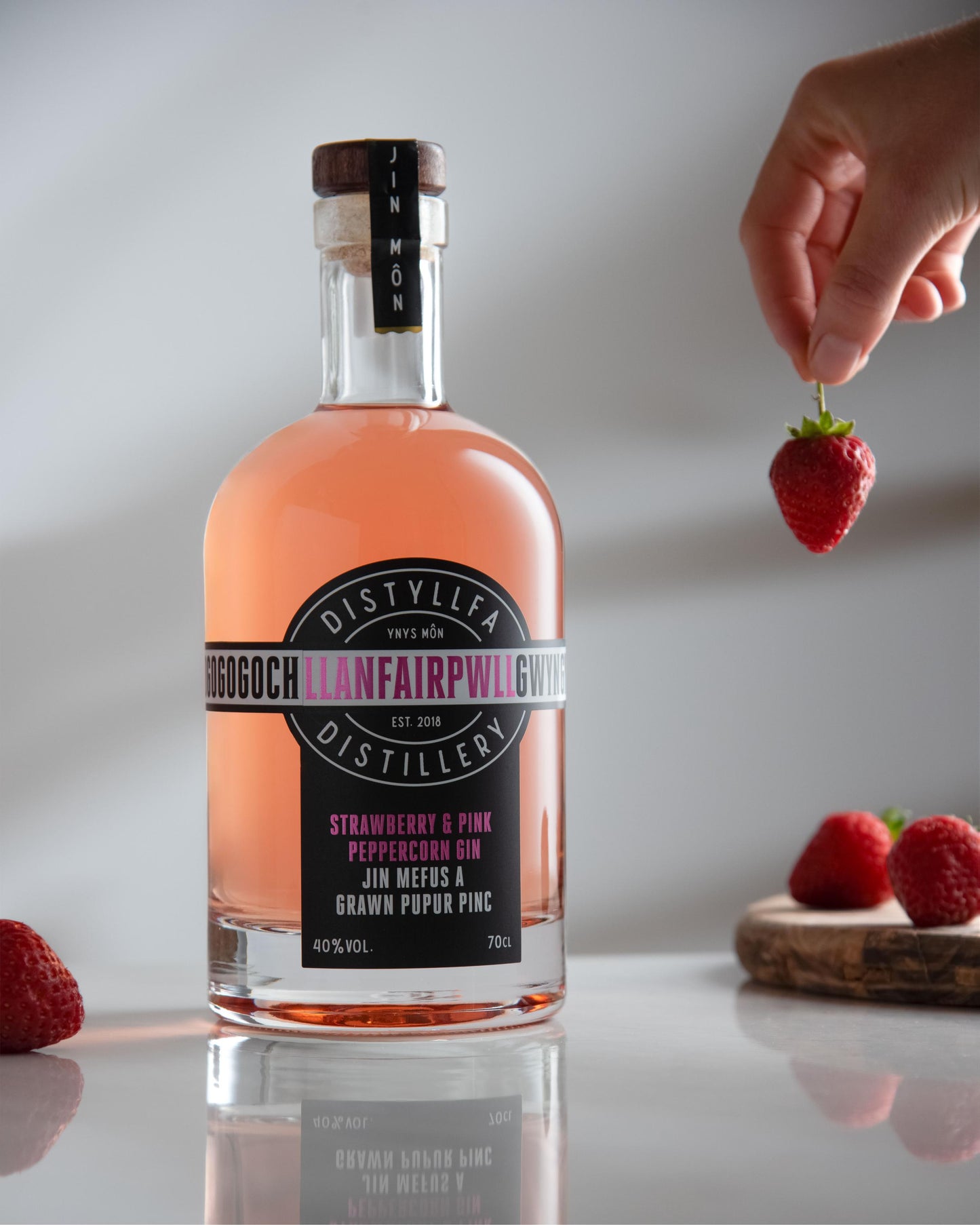 Strawberry & Pink Peppercorn Gin bottle with a hand holding a plump strawberry – Welsh craft gin from Anglesey with ripe strawberries and pink peppercorns